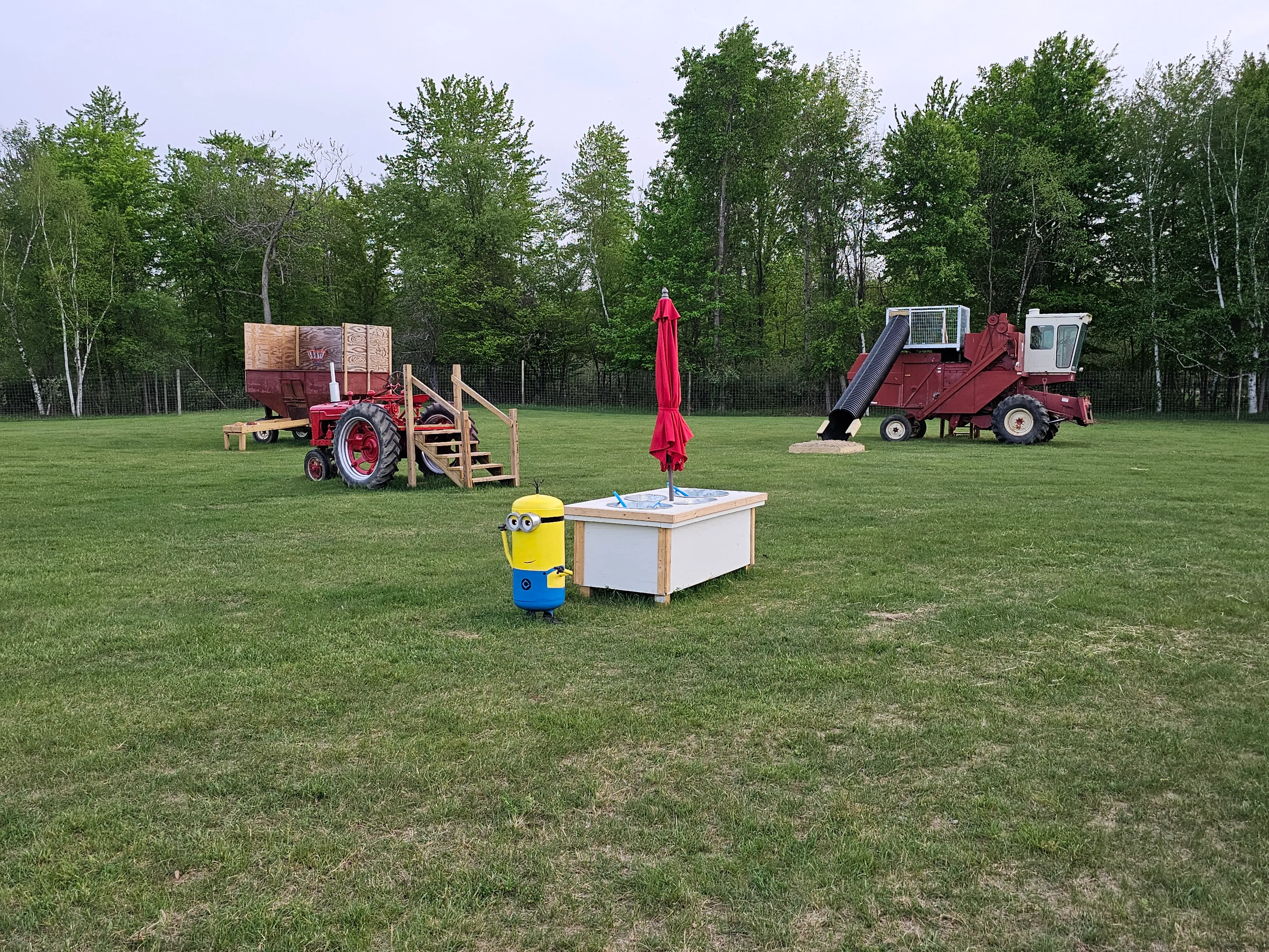 Wide view of the farm-equipment playground at Green's Deer Ranch — a retired wagon, vintage tractor, combine harvester, sandbox with umbrella, and open grass area